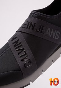 Svart sneaker med elastisk tygövre, med texturerade elastiska band med "CALVIN KLEIN JEANS"-logotyp. Tjock, vadderad vit sula.