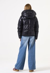 Zwarte pufferjack met capuchon, voorzien van een glanzende afwerking, gewatteerd ontwerp en elastische manchetten, gecombineerd met loszittende blauwe jeans.