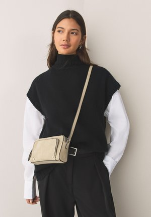 Nő fekete ujjatlan garbót visel fehér ing fölött, fekete nadrágot és bézs oldaltáskát, egyszínű háttér előtt állva.