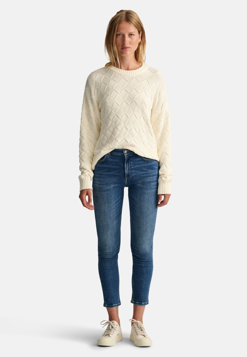 Krämfärgad kabelstickad tröja med rund hals, matchad med åtsittande blå denim jeans och ljusa sneakers.