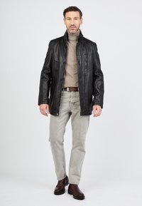 Schwarze Lederjacke mit Knopfverschluss und Seitentaschen, getragen über einem beigen Rollkragenpullover, kombiniert mit hellgrauen Jeans und braunen Stiefeln.