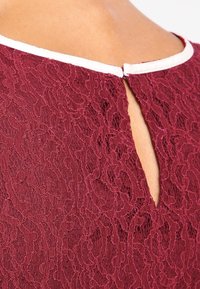 Haut en tissu en dentelle bourgogne avec un dos en trou de serrure et un col contrastant blanc. Présente des motifs floraux complexes et une surface texturée.