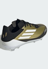 Botas de fútbol negras y doradas con una parte superior texturizada, un diseño elegante, tres bandas blancas y una suela de plástico transparente con tacos.