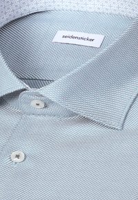 Col et boutons de chemise habillée bleu clair texturé avec une étiquette blanche indiquant "seidensticker" à l'intérieur du col.