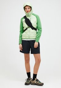 Giacca imbottita verde chiaro con cappuccio sopra una maglietta a maniche lunghe verde, abbinata a pantaloni corti neri e scarpe da trekking marroni. Borsa a tracolla nera.