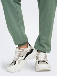 Baskets de sport au design robuste, composées de panneaux blancs, gris et noirs, semelle texturée et lacets blancs. Portées avec un pantalon de jogging vert.