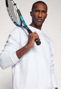 Sweatshirt blanc à manches longues avec des poignets côtelés, porté par un homme tenant une raquette de tennis bleue et noire, mettant en avant un design simple et détendu.