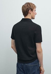 Černé polo tričko s krátkým límečkem, vyrobené z hladkého materiálu. Má krátké rukávy a elastický lem, v kombinaci s modrými denimovými kalhotami.