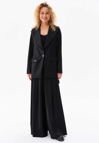 Zwarte blazer met een grote kraag, enkele knoopsluiting en twee voorzakken, gecombineerd met zwarte wijde broek met een zachte textuur.