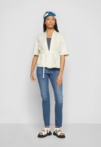 Lys beige innpakningsjakke, korte ermer, innsnevret midje med bånd. Kombinert med blå jeans og hvite sandaler med remmer. Blå mønstret hodepryd.