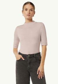 Helles rosa kurzärmeliges Rollkragenoberteil aus einem weichen Strickstoff. Hat eine figurbetonte Silhouette und gerippte Bündchen, kombiniert mit dunklen, hochgeschnittenen Jeans.