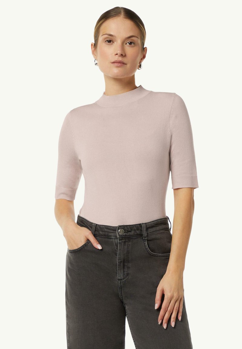 Helles rosa kurzärmeliges Rollkragenoberteil aus einem weichen Strickstoff. Hat eine figurbetonte Silhouette und gerippte Bündchen, kombiniert mit dunklen, hochgeschnittenen Jeans.