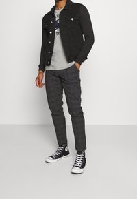 Giacca di denim nera con bottoni d'argento, T-shirt grigia con stampa, pantaloni grigi a quadri e sneakers alte nere con dettagli bianchi.