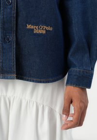 Donkerblauwe denimshirt met oranje stiksels en "Marc O'Polo DENIM" geborduurd in het oranje boven een witte gerimpelde rok, met een ontspannen hand te zien.