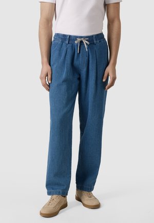 Mann trägt locker sitzende blaue Jeanshose mit elastischem Bund und grauem Kordelzug, weißes Hemd und beigen Sneakers, steht vor einem einfarbigen Hintergrund.