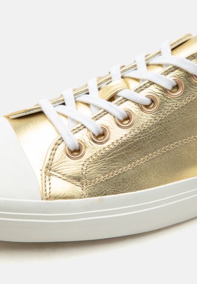 Εγγύς πλάνο χρυσού μεταλλικού αθλητικού παπουτσιού με λευκά κορδόνια, χρυσές οπές και λευκή λαστιχένια σόλα σε απλό φόντο.