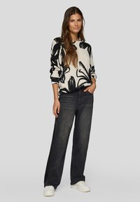 Cremefarbener Pullover mit schwarzem Blumenmuster, kombiniert mit dunkelblauen weit geschnittenen Jeans und weißen Sneakers. Langes Haar lässig gestylt, steht vor einem neutralen Hintergrund.