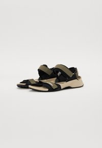 Deux sandales avec un design noir et vert olive, dotées de sangles ajustables et d'une semelle beige clair. Semelle extérieure en caoutchouc texturé pour une meilleure adhérence.