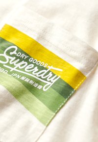 Parche de tela blanca con rayas horizontales verdes y amarillas. El texto dice "DRY GOODS" y "Superdry". Textura suave con bordes cosidos.