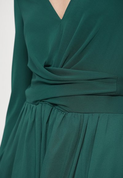 Πράσινο φόρεμα με βαθύ V-λαιμόκοψη, σφηκοφωλιά στη μέση και αέρινη πλισέ φούστα από ελαφρύ ύφασμα με λεία υφή.
