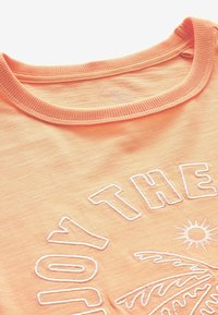 Tissu couleur pêche avec un col rond côtelé et un texte brodé blanc ainsi que des motifs de soleil et de feuille de palmier.