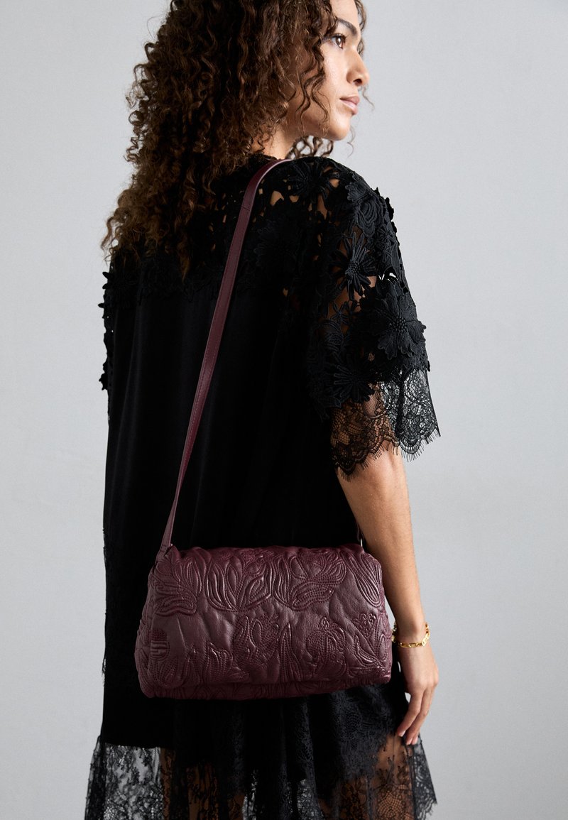 Burgundy leather shoulder bag with embossed floral design, elongated shape, and a single strap, paired with a black lace top.