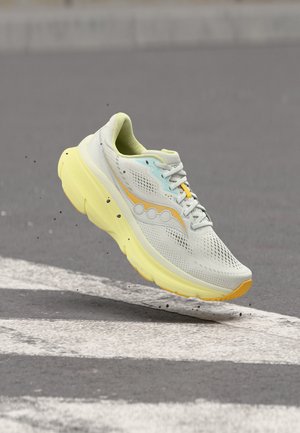 Lys grå og gul løpesko med tykk såle, svevende over asfaltvei med hvite fotgjengerovergangslinjer, små partikler flytende i nærheten.