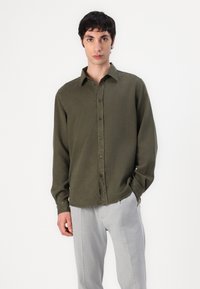 Olive green long-sleeve shirt with a button-down collar, front placket, and soft texture, paired with light grey trousers.