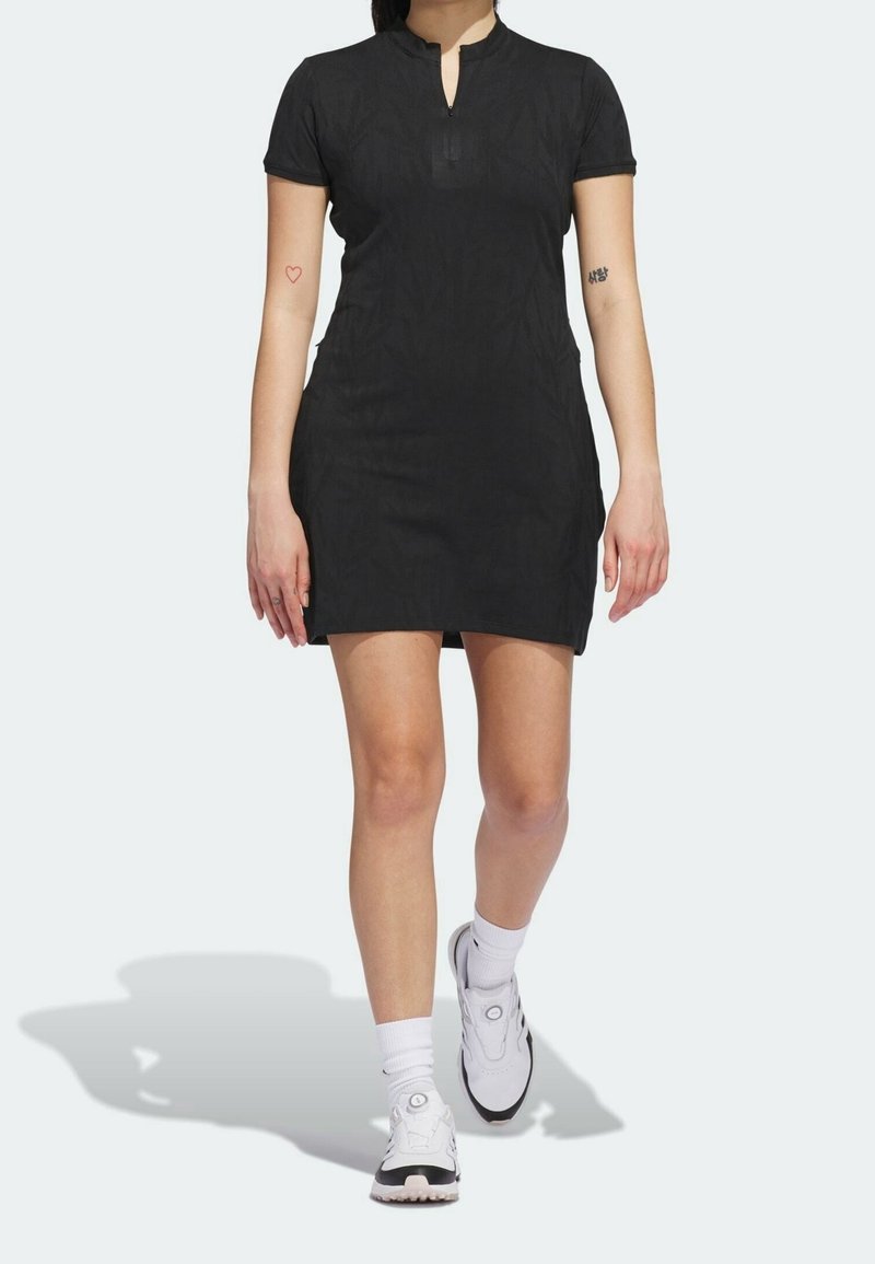 Femme portant une robe courte noire avec un décolleté en forme de trou de serrure, des chaussettes blanches, des baskets blanches et noires, et des tatouages sur les deux bras supérieurs.