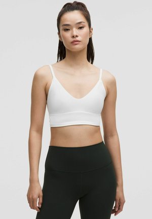 Giovane donna con capelli scuri e lunghi, indossa un reggiseno sportivo bianco e leggings neri a vita alta su uno sfondo chiaro e semplice.