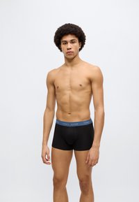 Μαύρα βαμβακερά βαμβακερά εσώρουχα τύπου μπόξερ με μπλε ελαστική μέση που φέρει το λογότυπο "TOMMY HILFIGER". Το μοντέλο στέκεται μπροστά σε ένα απλό φωτεινό φόντο.