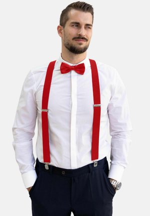 Rote Hosenträger mit silbernen Clips, getragen über einem weißen Hemd, kombiniert mit einer roten Fliege. Die Person hat dunkles Haar und einen Bart.