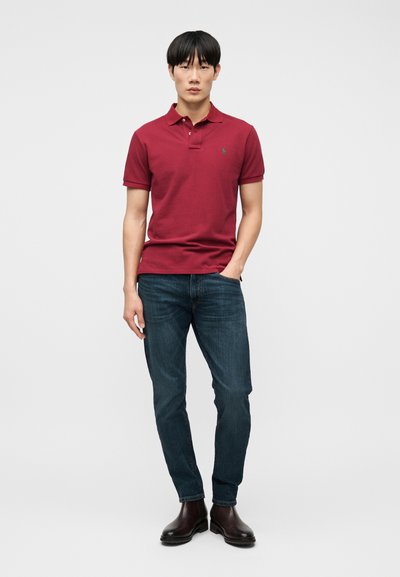 Νέος άνδρας που στέκεται, φοράει κόκκινο πόλο μπλουζάκι, σκούρα μπλε τζιν και σκούρα καφέ μπότες, μπροστά σε απλό ανοιχτό γκρι φόντο.