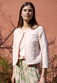Hellrosa, strukturierte, cropped Jacke mit rundem Ausschnitt und vorderer Tasche, kombiniert mit einer pastellfarbigen Bluse und einem rock mit blattförmigem Muster.