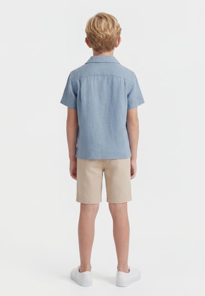 Enfant blond de dos, portant une chemise à manches courtes bleu clair, un short beige et des baskets blanches sur un fond uni.