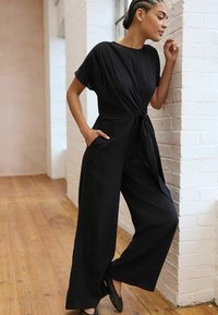 Schwarzer Jumpsuit mit kurzen Ärmeln und geknotetem Bund, aus leichtem Stoff. Er hat weite Beine und zwei Fronttaschen.