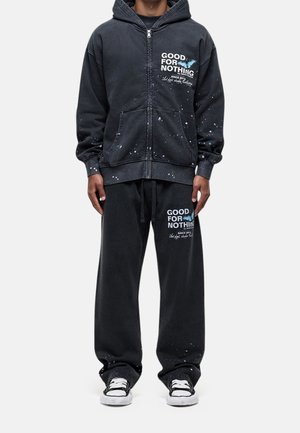 Personne portant un sweat à capuche noir zippé et un pantalon ample assorti avec le texte « GOOD FOR NOTHING » et des motifs de papillons bleus.
