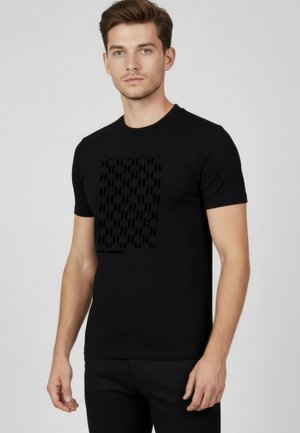 Jeune homme aux cheveux châtain clair portant un t-shirt noir à manches courtes avec un motif répétitif subtil et le texte "Karl Lagerfeld".