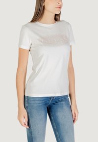 Camiseta blanca de manga corta con un logotipo "GUESS" texturizado en acentos de cristal en la parte delantera; se ajusta al cuerpo con un escote redondo.