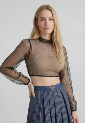 Blonde Frau mit langen Haaren trägt ein transparentes schwarzes Crop-Top mit Punkten und einen hoch taillierten blauen plissierten Rock mit dekorativer Gürtelschnalle.