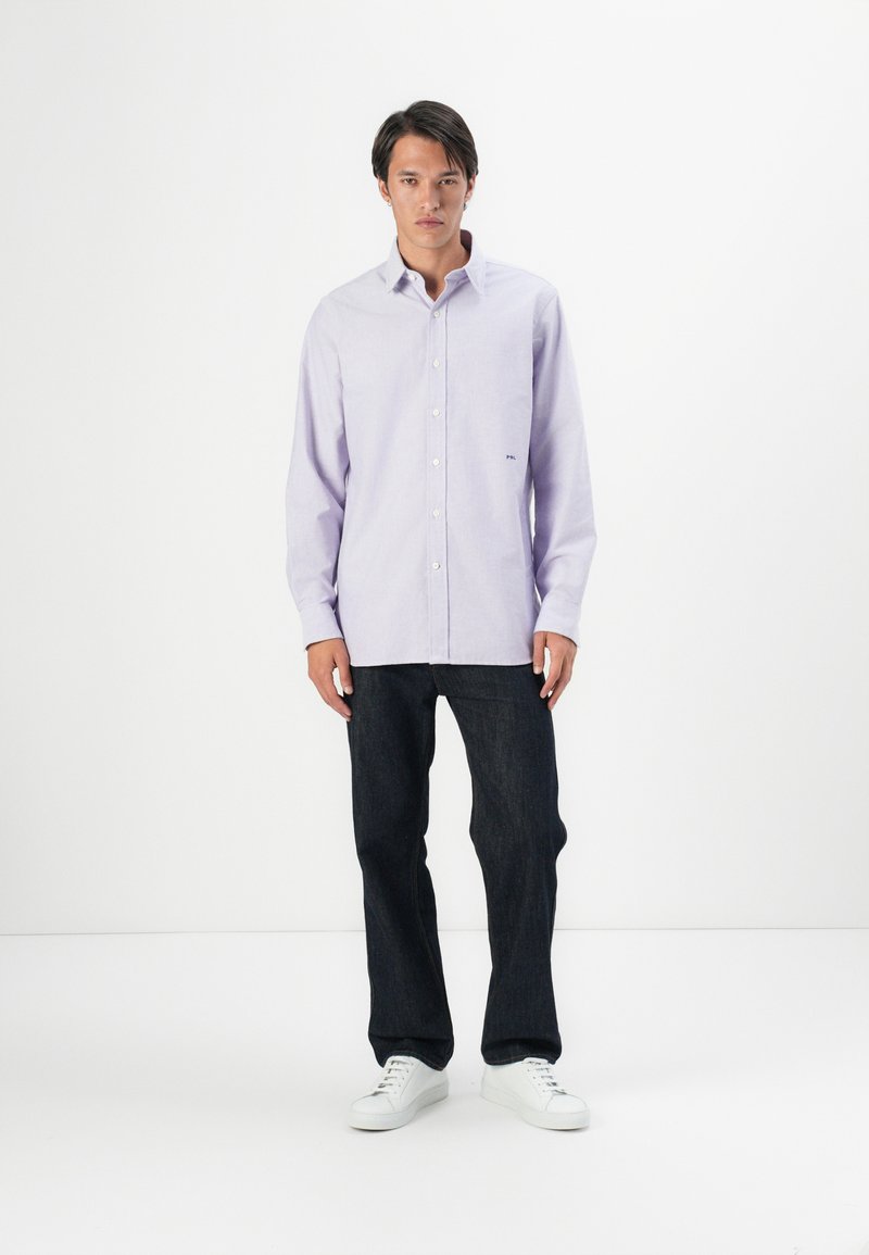 Camisa de botón de manga larga de color púrpura claro con cuello y un pequeño logo bordado en el pecho, combinada con jeans oscuros y zapatillas blancas.