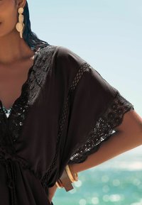 Kaftan noir avec dentelle, coupe ample, tissu texturé et taille cintrée. Porté avec de grandes boucles d'oreilles décoratives et des bracelets en bois.