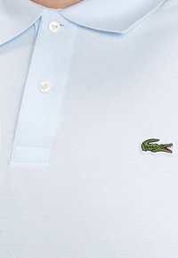 Světle modré polo tričko se dvěma bílými knoflíky a malým zeleným krokodýlem na prsou.