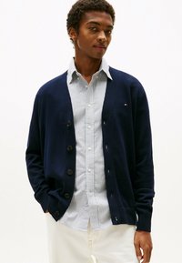 Marineblauer Cardigan mit Knopfverschluss, strukturiertem Stoff und gerippten Bündchen, über einem weiß-blau gestreiften Hemd getragen. Das Model posiert vor einem neutralen Hintergrund.