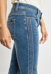 Detailní záběr na osobu v modrých denimových džínách s trojitým vertikálním švem podél strany a viditelnou zadní kapsou, ruce uvolněně po stranách.