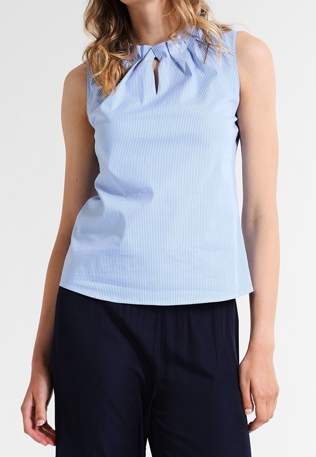 Femme portant un haut sans manches rayé bleu clair avec un col froncé et un détail en forme de goutte, associé à un pantalon bleu marine foncé.