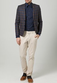 Brun rutete blazer over en marineblå skjorte, kombinert med lyse beige bukser og brune sko. Blazeren har en smal rev og lommer.