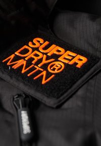 Czarna tkanina kurtki z teksturowaną naszywką z napisem "SUPER DRY" w wyraźnych pomarańczowych literach. Zawiera czarny akcent w postaci zamka.