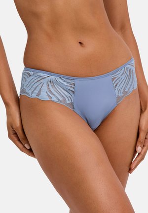 Donna che indossa biancheria intima stile bikini azzurro chiaro con pannelli laterali in pizzo con motivo a foglie su sfondo chiaro.