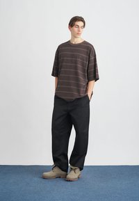 Camiseta de gran tamaño a rayas marrón, pantalones sueltos negros, botas grises con suelas de goma. Modelo de pie sobre una alfombra azul frente a un fondo blanco.